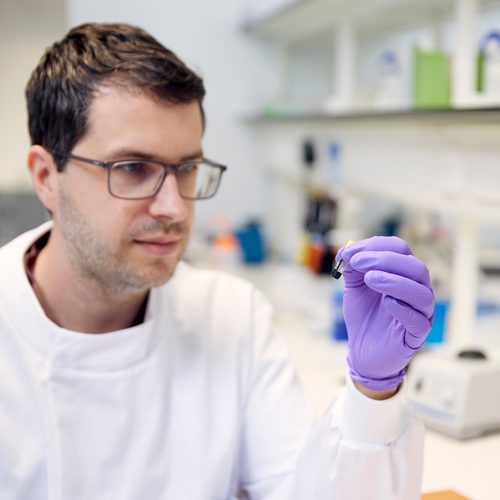 Scientist in lab coat holding and staring at sample in a microtube.